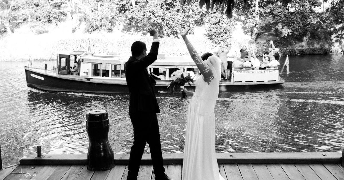Bride and groom raising their arms, facing a canal boat with cheering passengers at Amora Herencia Riverwalk Melbourne