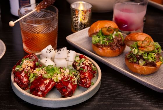 An appetizing spread of small plates and drinks, featuring savory sliders, glazed chicken wings, and two cocktails