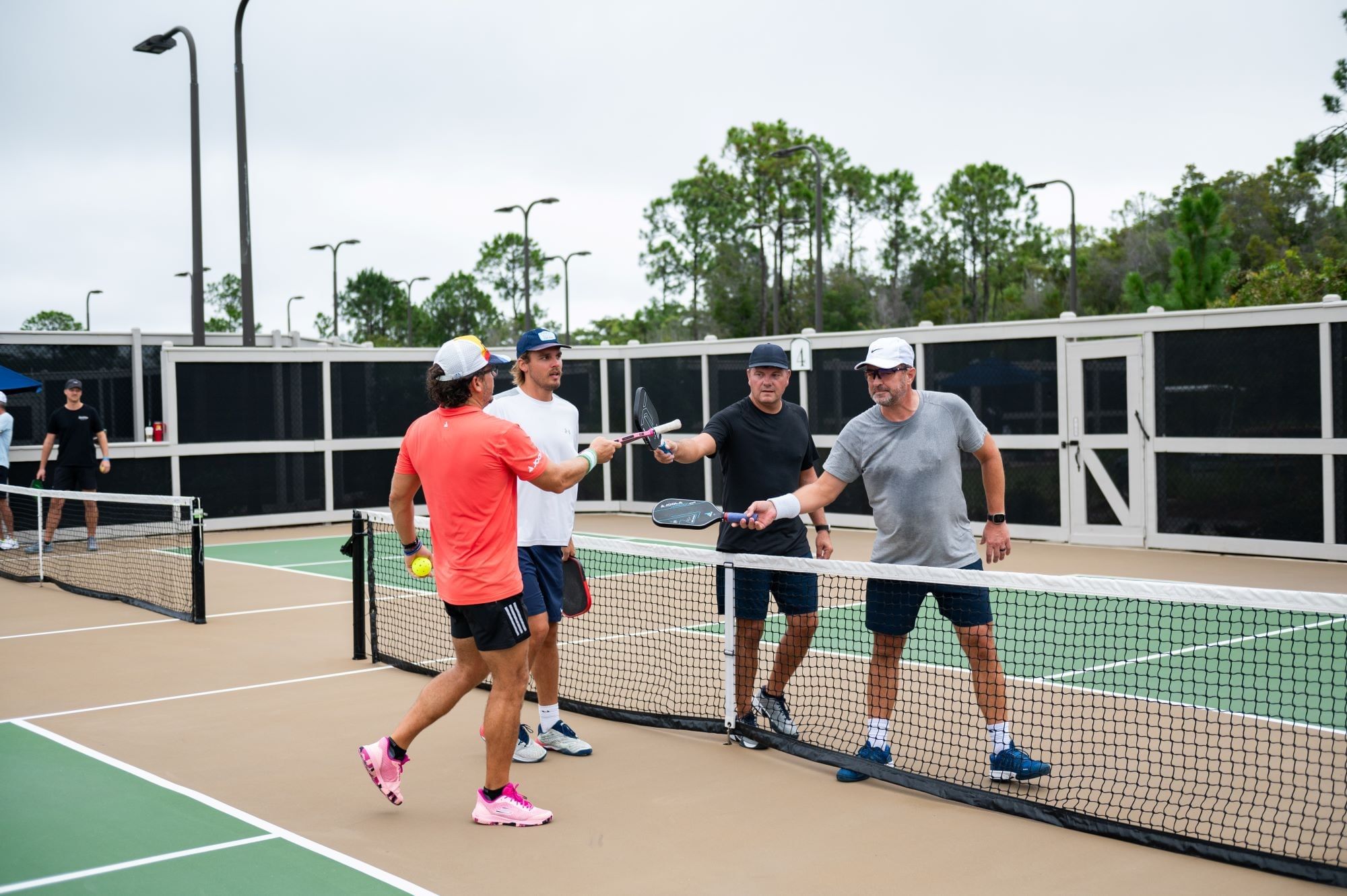 Four men on a pickleball court with two holding paddles, one holding a ball, and trees in the background.