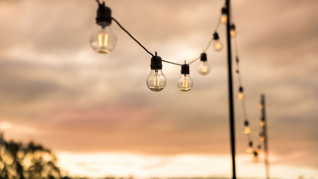 String of glowing light bulbs against a cloudy sunset sky with trees in the background.