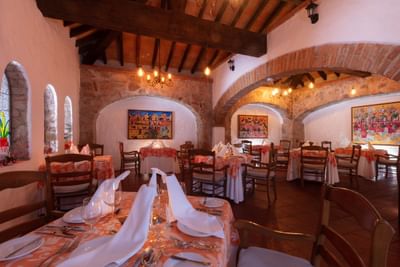 A rustic dining room with set tables, brick arches, wooden beams, and vibrant artwork on the walls.