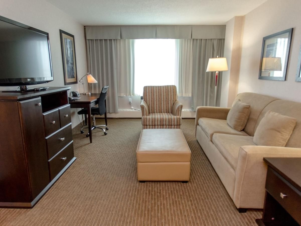 Hotel suite living area with a sofa, armchair, ottoman, TV and work desk by the window.