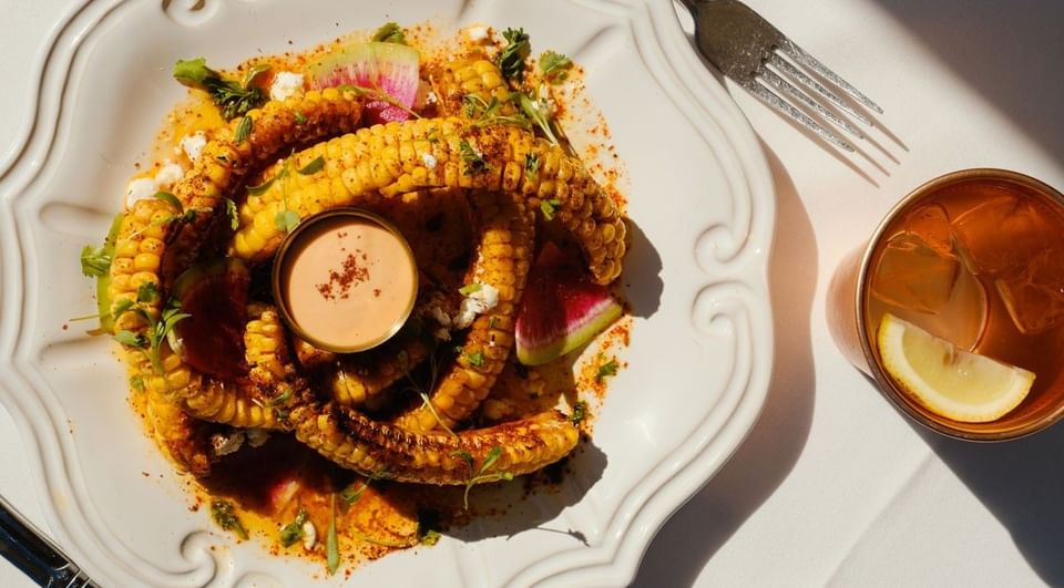 Grilled corn ribs with dipping sauce on a white ceramic plate, and an iced drink served under a bright sun at Petit Ermitage