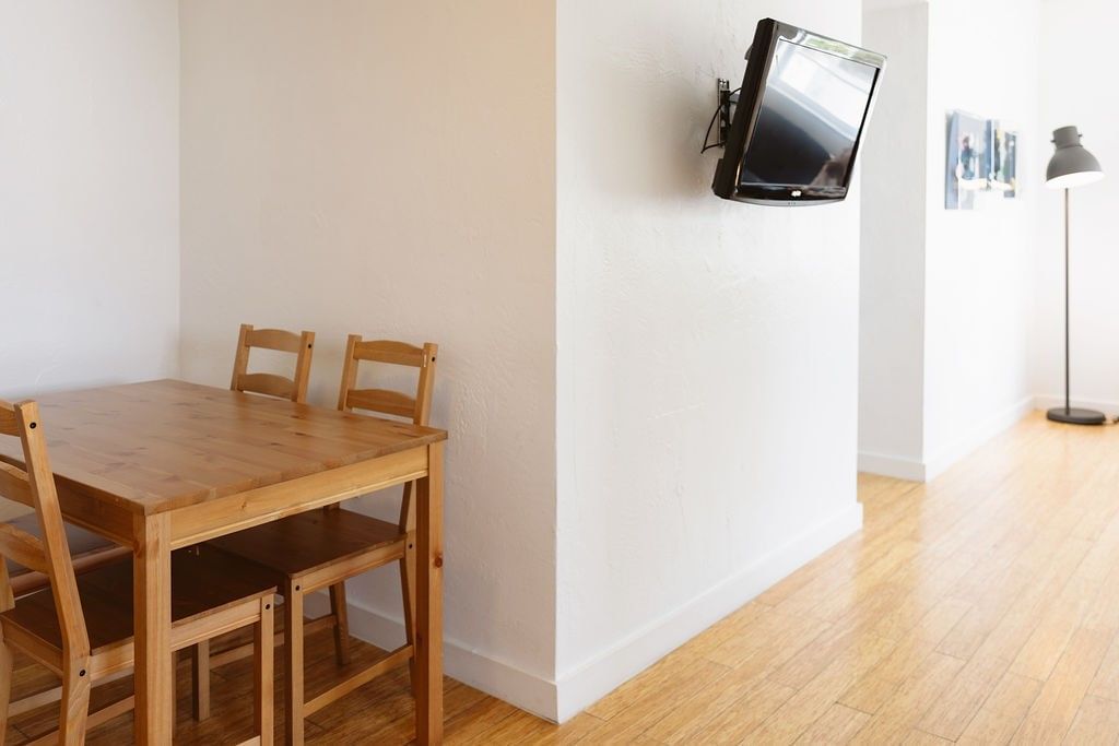 A small dining area with a table and chairs, a wall-mounted TV, and a floor lamp in the background.