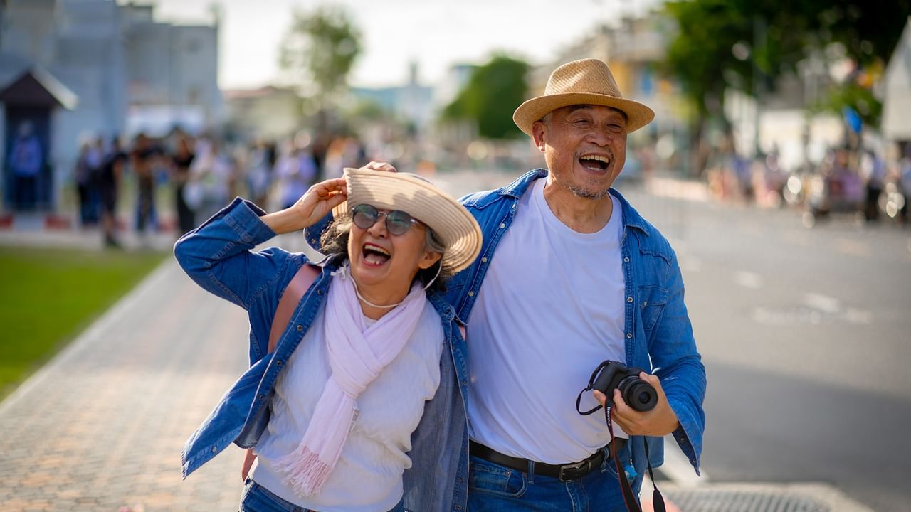 Two elderly people laughing and walking on a sidewalk with a crowd in the background.
