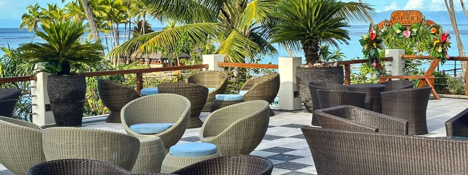 Round lounge chairs by a dark wicker sofa under palm trees in Sunset Terrace Bar at Warwick Fiji Resort and Spa