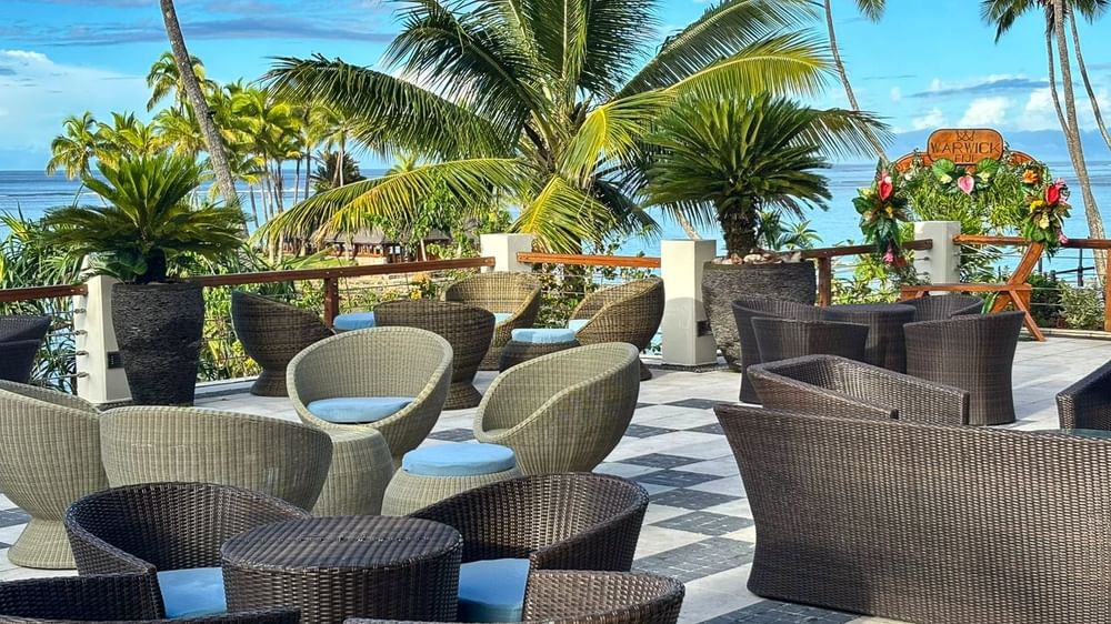 Round lounge chairs by a dark wicker sofa under palm trees in Sunset Terrace Bar at Warwick Fiji Resort and Spa