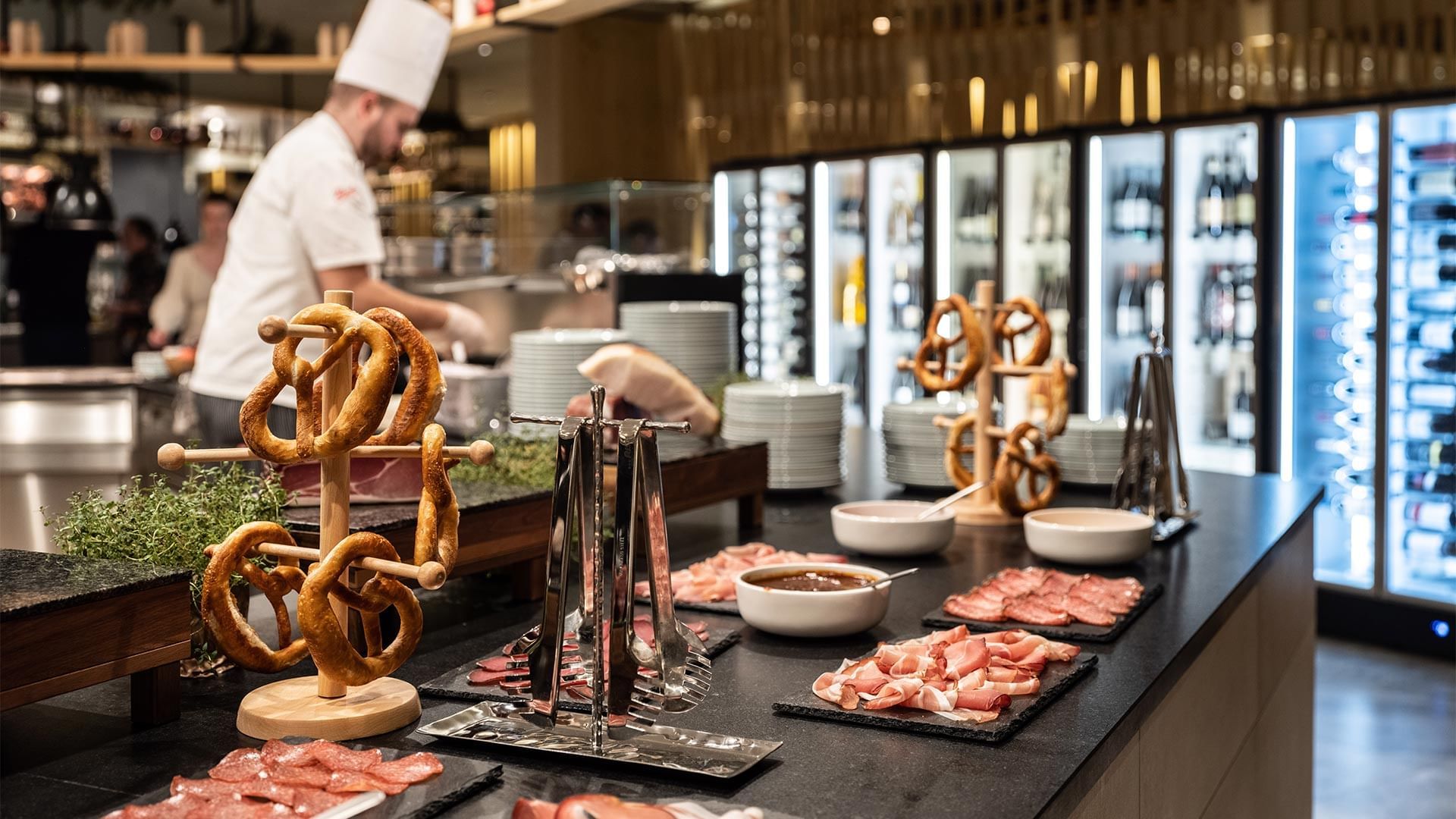 Küchenchef in weißer Uniform bereitet Speisen hinter einer Theke mit Wurstplatten und Brezeln zu.