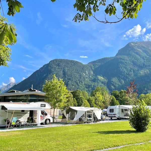 Diversi camper e tende parcheggiati su un prato verde con montagne sullo sfondo.
