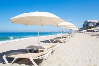 White beach umbrellas and lounge chairs line a sandy shore with ocean waves and coastal buildings at Playa Los Arcos Puerto Vallarta