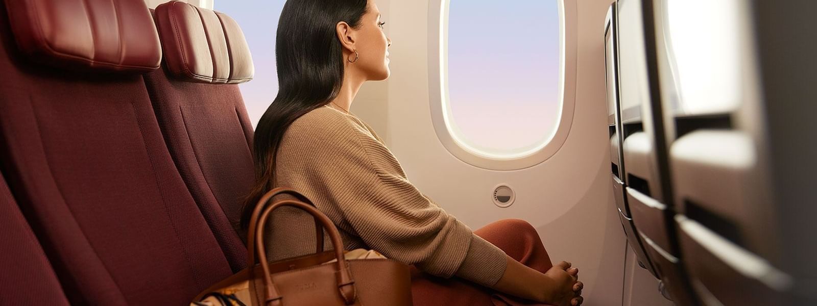 A woman sits in a maroon airplane seat, gazing out the window with a brown handbag by her side.