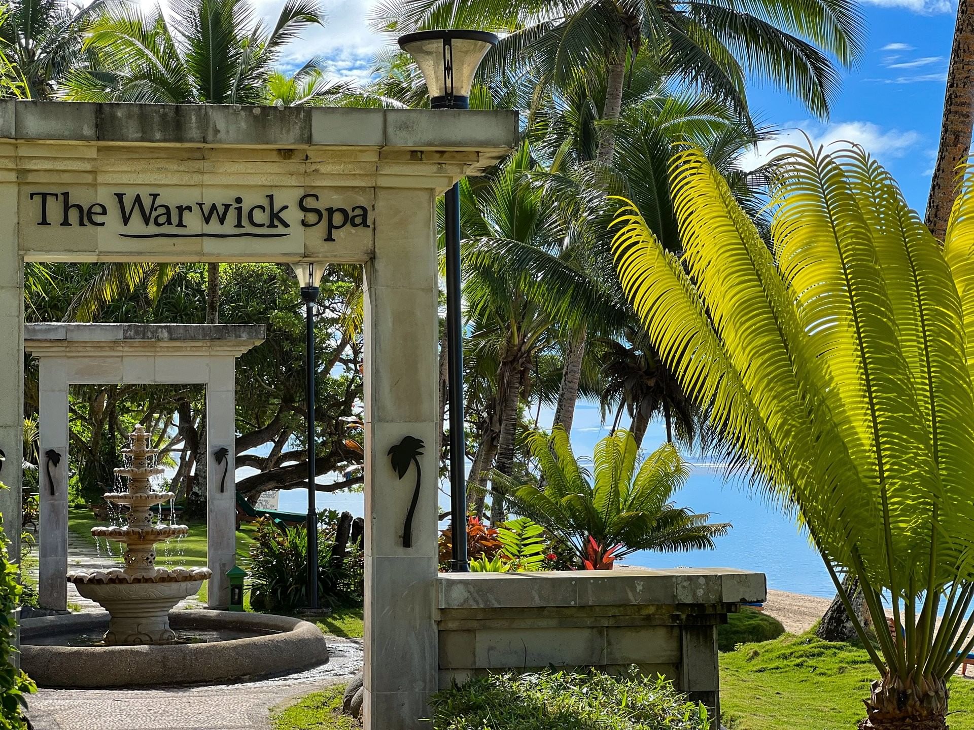 Entrance of Warwick Spa Stone archway with spa signage by a tiered fountain under palm trees at Warwick Fiji Resort and Spa