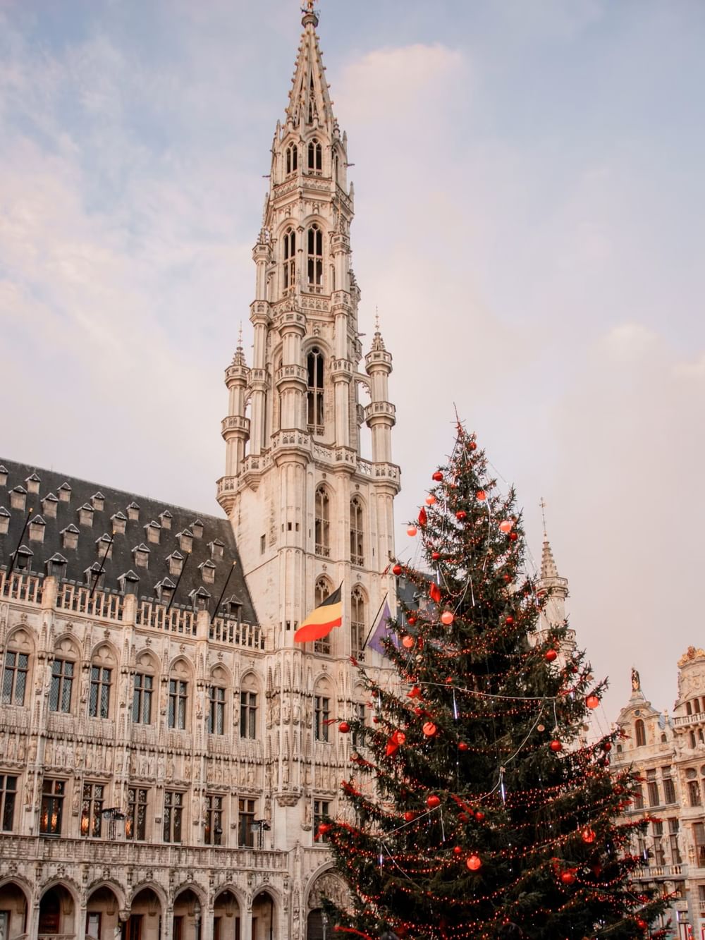 Ornate Gothic bell tower rises above a decorated Christmas tree and historic buildings in Brussels' Grand Place square.