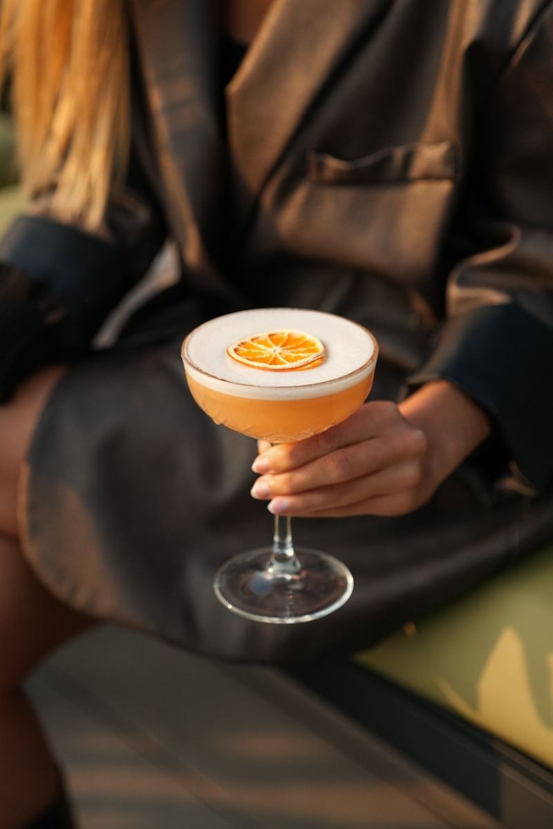 Hand holding a cocktail in a glass by a dried orange slice under soft light at Warwick Grand Place Brussels