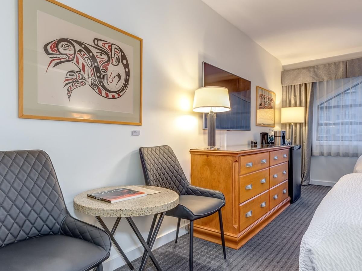 A hotel room with two beds, a table, two chairs, a dresser, and a framed Indigenous art piece.