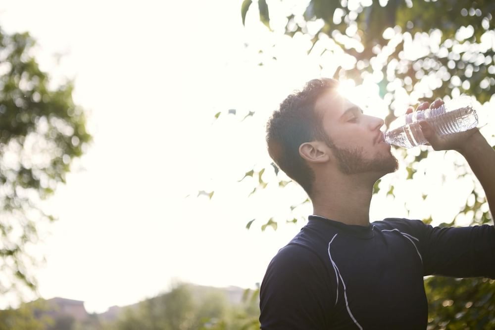 A man wearing a black shirt drinking a water bottle in nature as the sun gleams in the background.