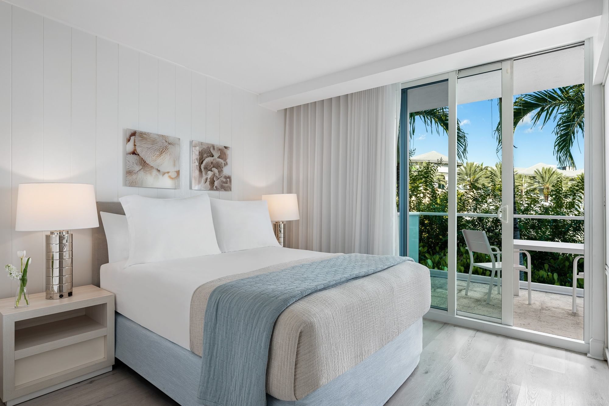 A hotel room with a large bed, two lamps, and a balcony with a view of palm trees