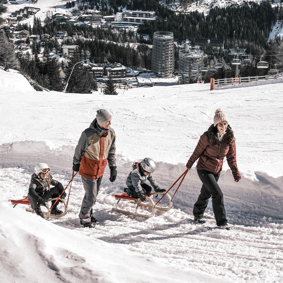 A family of four pulling two children on sleds down a snowy slope with buildings in the background.