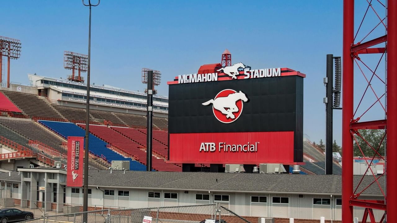 Exterior view of McMahon Stadium featuring large scoreboard with team logo and colorful seating sections under blue sky.
