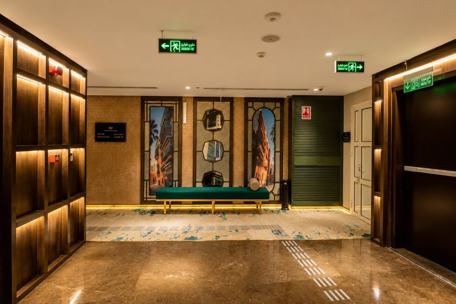 Teal bench under decorative wall mirrors in elevator lobby with emergency exit signs at Naviti by Warwick Jeddah