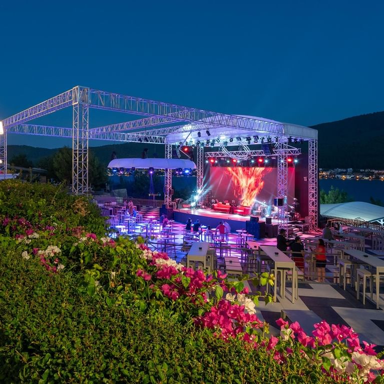 Открытая вечерняя площадка для мероприятий с сценой, освещением и зоной для зрителей. Уличные стулья и сцена, подготовленные для мероприятия в Titanic Luxury Collection Bodrum.