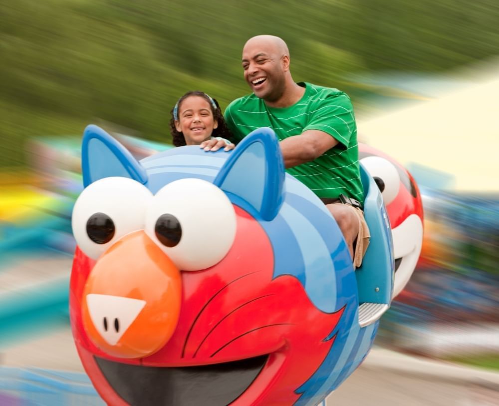 A father and young daughter smile while riding in a ride car designed to look like Elmo dressed as a cat.