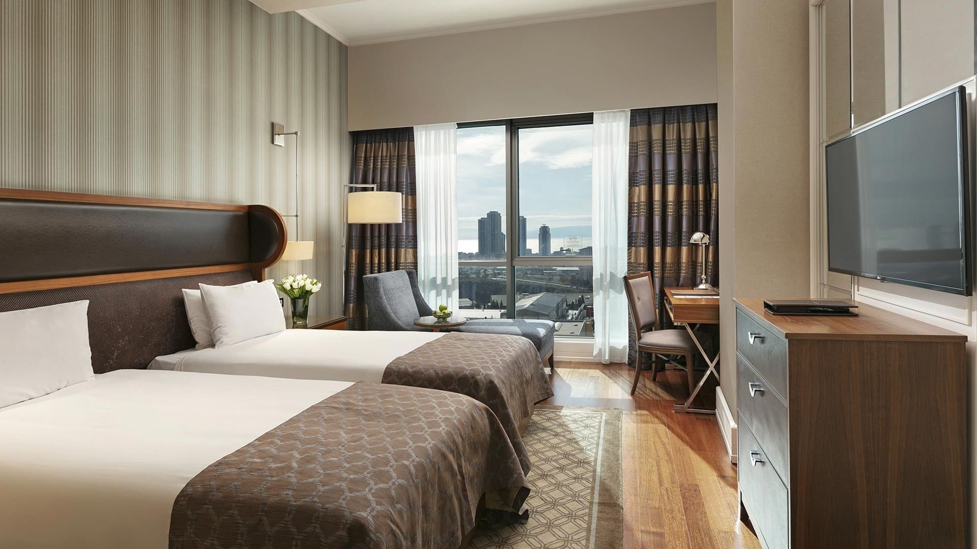 Twin beds, cozy lounger, TV, and work desk with a balcony city view in Superior Room at Titanic Business Kartal