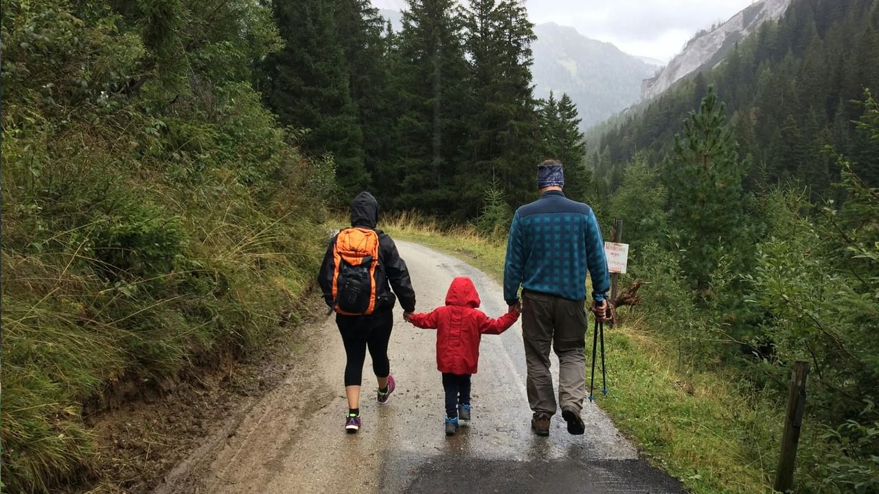 Two adults and a child hike on a wet mountain trail with backpacks and hiking poles.