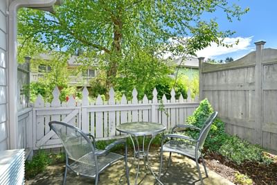 Patio with two chairs and table near a white picket fence and green wooden fence under a blue sky.