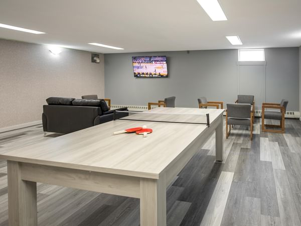 A room with a ping pong table, chairs, and a TV showing a basketball game.