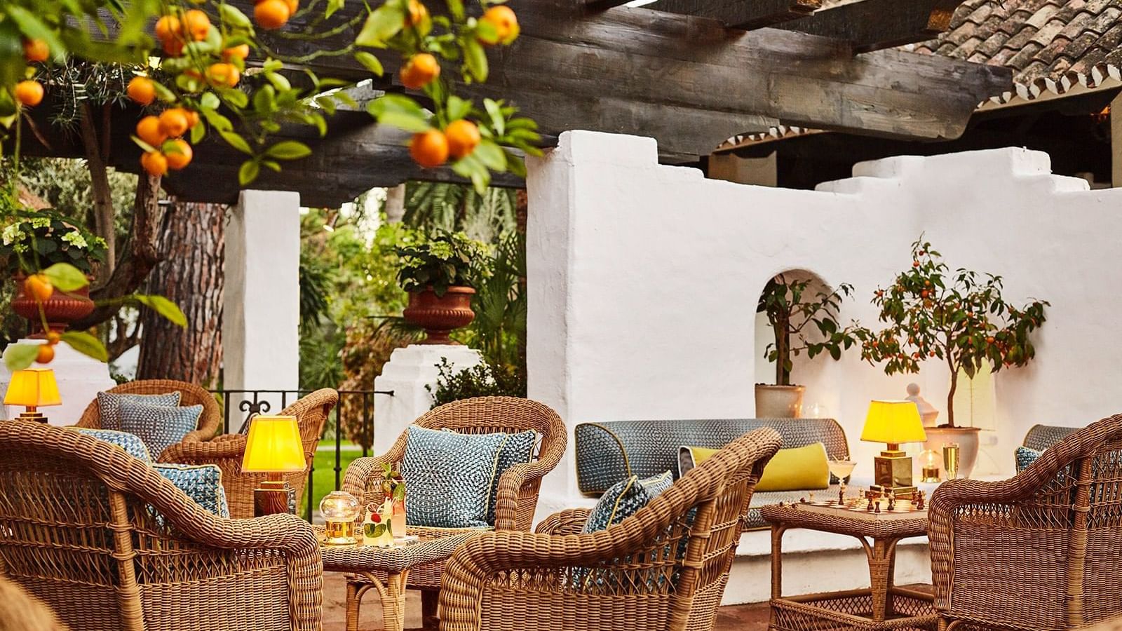 Wicker chairs and tables placed near a white stucco wall, under an orange tree and a wooden pergola at Marbella Club