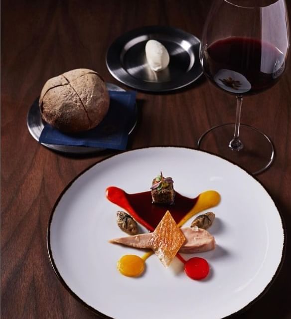 Fine dining plate featuring a gourmet dish with various colorful elements, accompanied by a glass of red wine