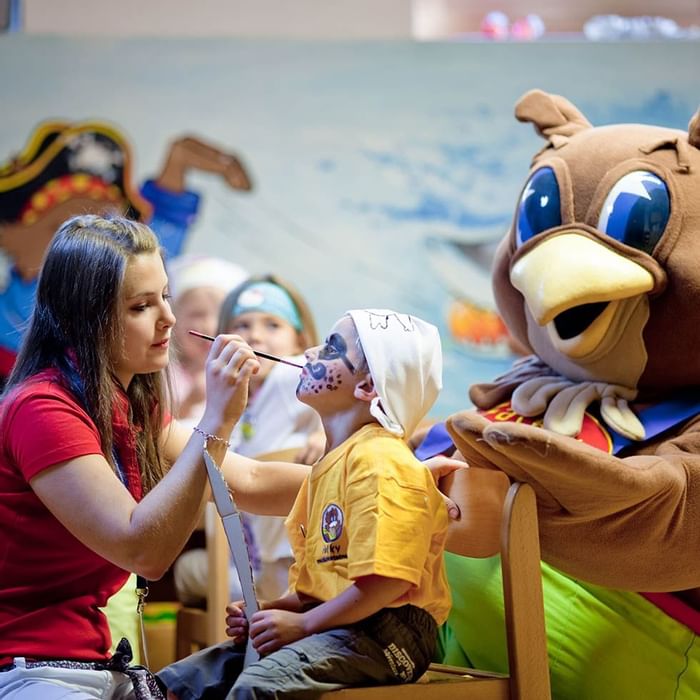 Child with face paint sitting next to a mascot at Falkensteiner Family Resort Lido