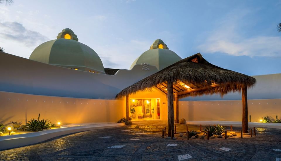 Entrance to the Quinta Real Huatulco under white domes by a thatched hut surrounded by desert plants at night