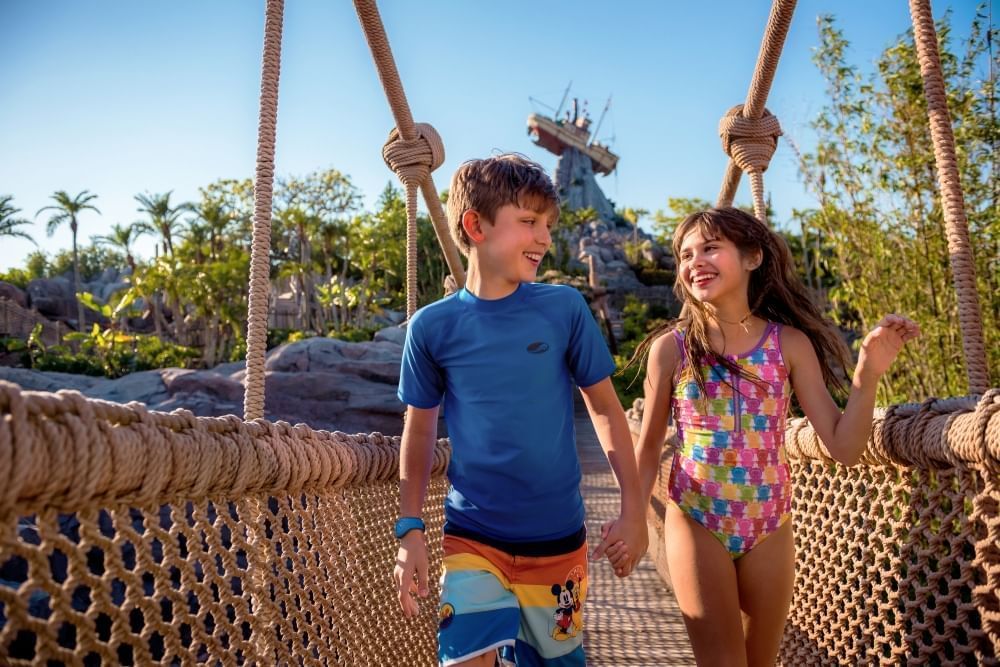 A boy and girl in bathing suits holding hands cross a rope bridge with a marooned fishing boat perched on top of a mountain in the distance.