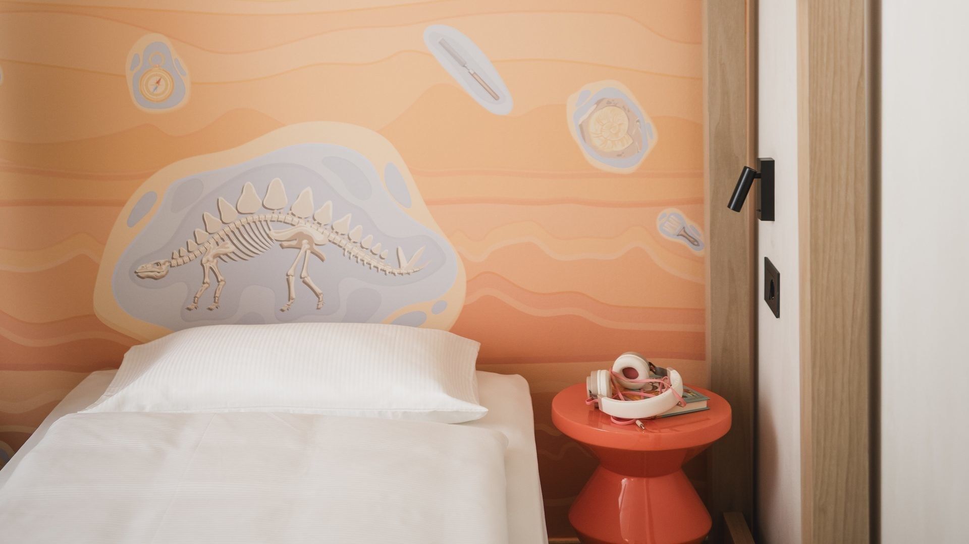 Camera da letto con dinosauro dipinto sulla parete, letto singolo e tavolino rosso.