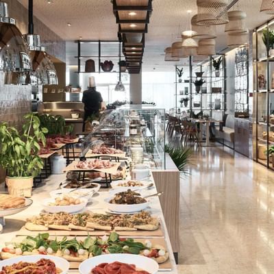 Long buffet table with various food dishes, a person in the background, and shelves with plants.