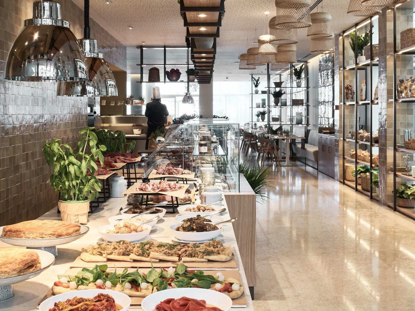 Lungo buffet in un ristorante con piatti di carne, pesce, verdure e pane, sotto luci sospese.