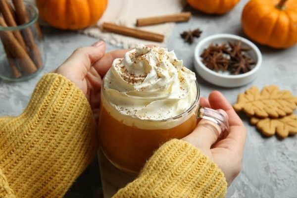 A pair of hands wraps around a pumpkin spice coffee topped with whipped cream and surrounded by fall spices and pumpkins.