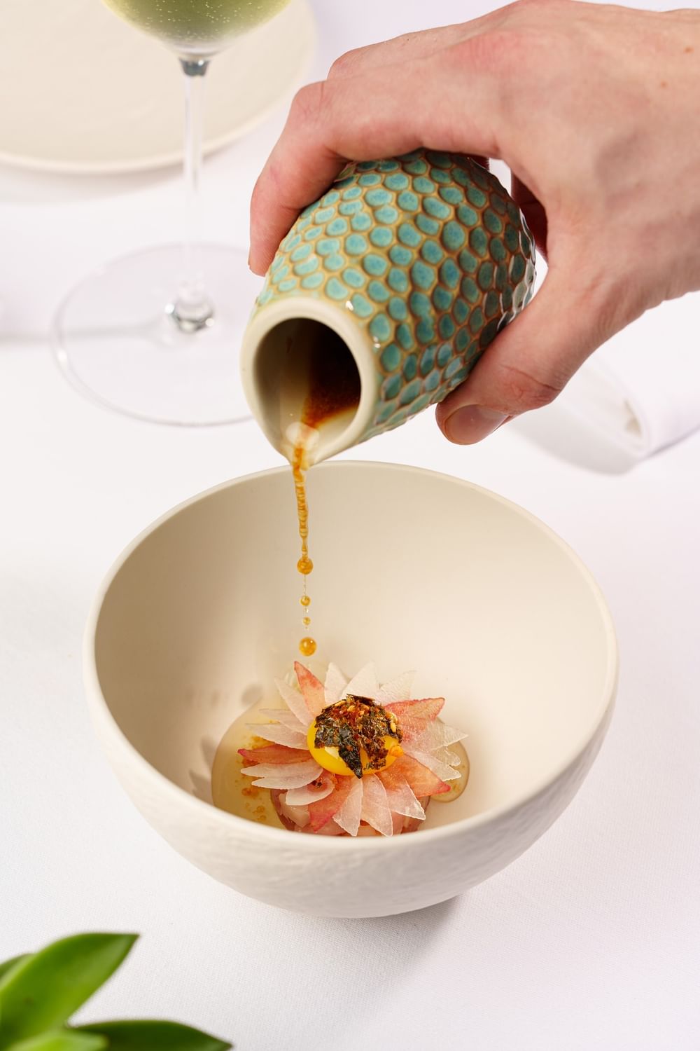Hand pouring sauce from a textured jug by a bowl with an edible flower at The Capital Hotel, Apartments and Townhouse