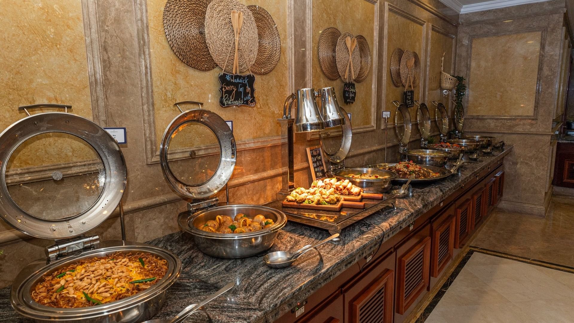 Stainless steel chafing dishes by woven wall decor under warm spotlights in L’auberge at Warwick Doha Hotel