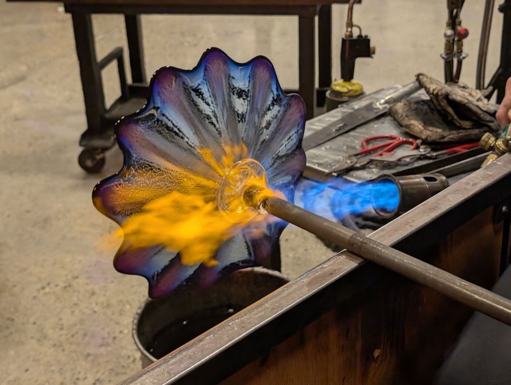 Wavy glass piece shaped by a blue flame on a metal rod under a warm glow at Warwick Seattle