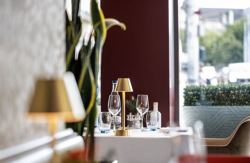 Gold lamp by wine glasses on a white table under a tall plant near a window in Téséo at Warwick Geneva