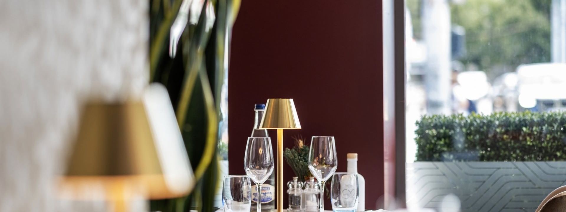 Gold lamp by wine glasses on a white table under a tall plant near a window in Téséo at Warwick Geneva