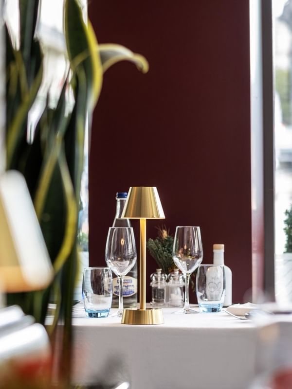 Gold lamp by wine glasses on a white table under a tall plant near a window in Téséo at Warwick Geneva