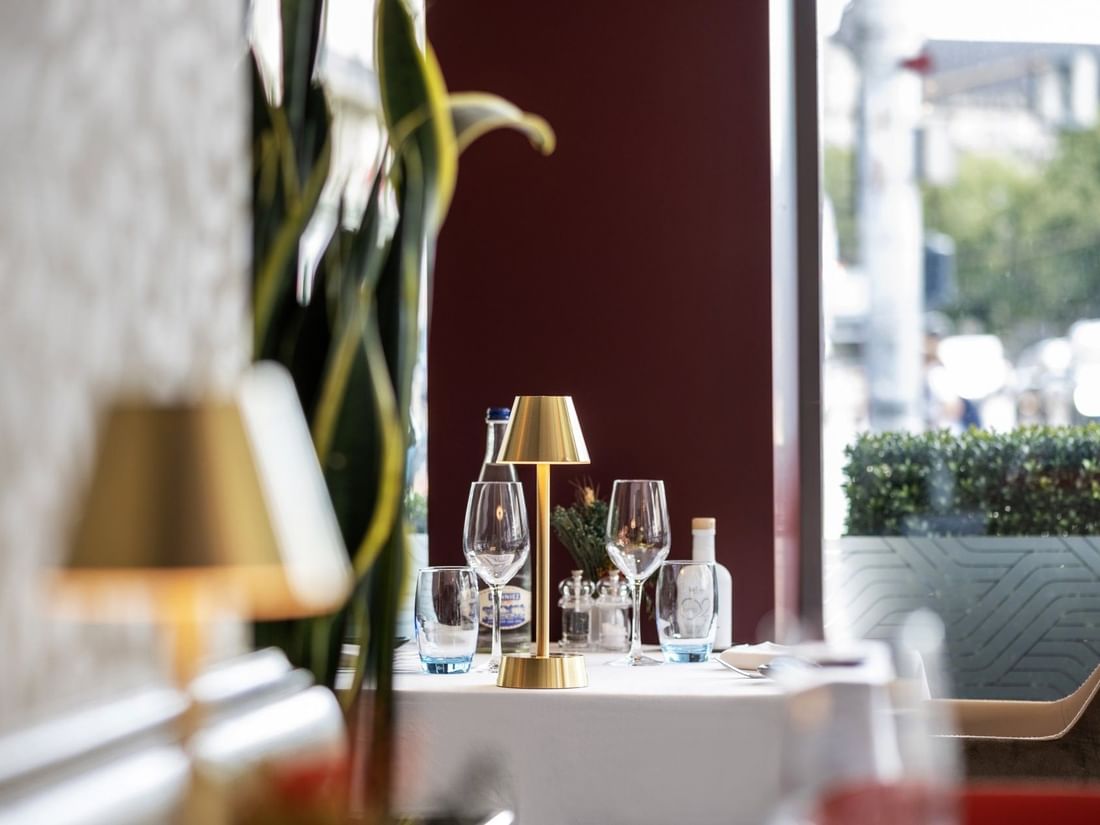 Gold lamp by wine glasses on a white table under a tall plant near a window in Téséo at Warwick Geneva