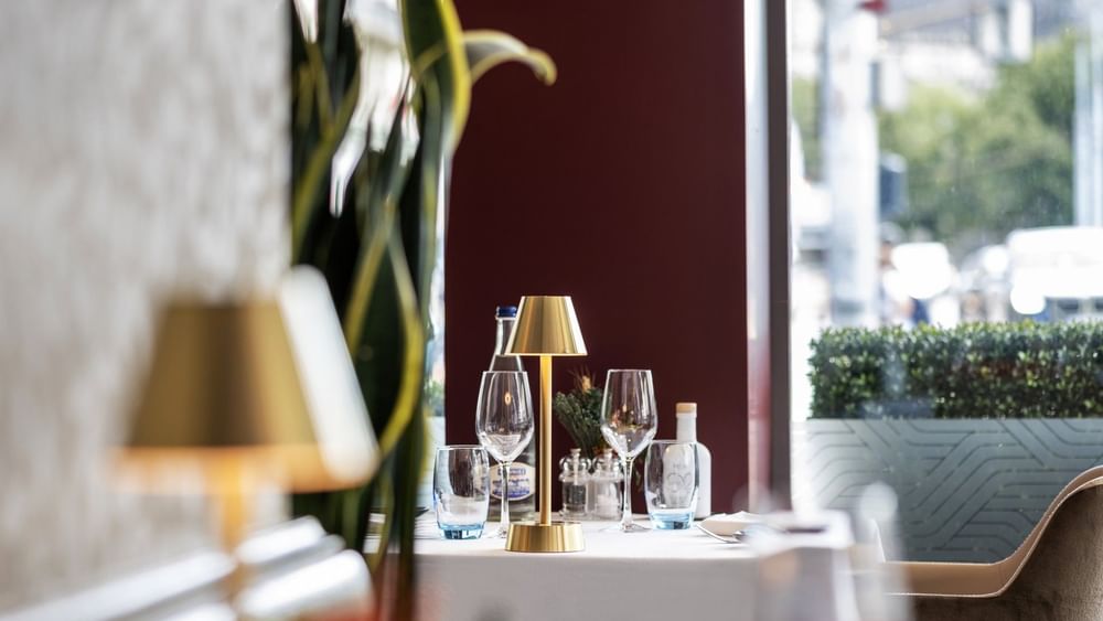 Gold lamp by wine glasses on a white table under a tall plant near a window in Téséo at Warwick Geneva