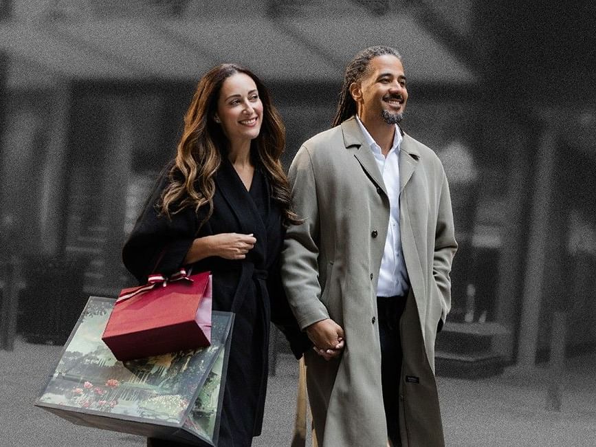 Couple walking down a street, woman holding shopping bags, man in trench coat holding her hand.