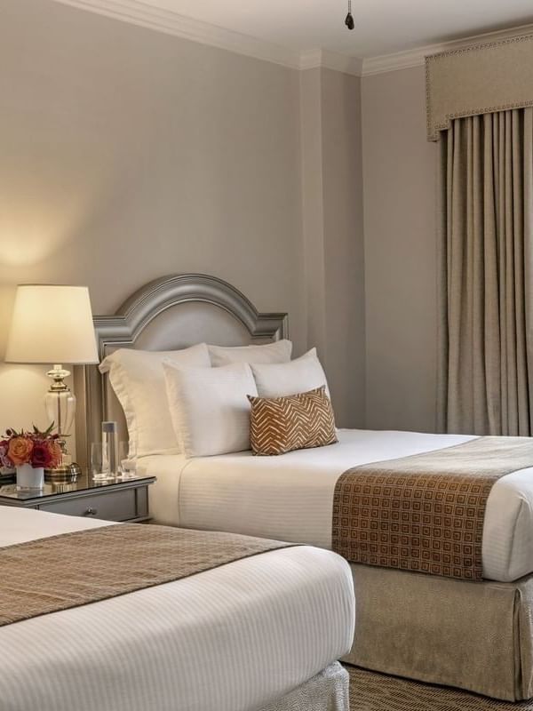 Two double beds with tan runners by a shared nightstand under a glowing lamp in Deluxe Double at Warwick Melrose Dallas