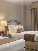 Two double beds with tan runners by a shared nightstand under a glowing lamp in Deluxe Double at Warwick Melrose Dallas